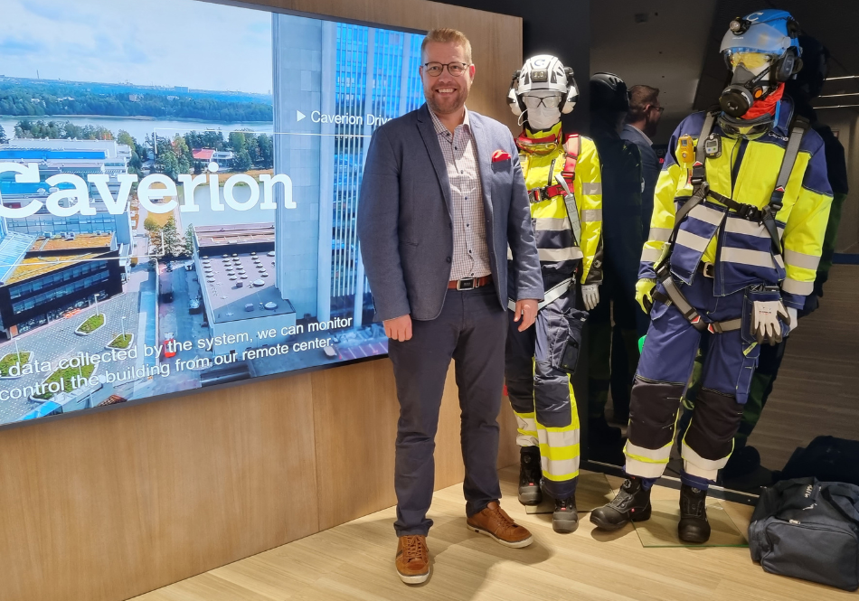 Jonni Moislahti seisoo pukupäällä Caverionin showroomissa. Hänen vasemmalla puolella on iso tv, jossa on teksti Caverion ja oikealla puolella on kaksi mallinukkea työvaatteissa.
