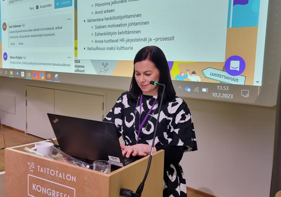 Minna Pohjola pitää Taitotalon strategiapäivässä puheenvuoroa voittavasta kulttuurista. Minna seisoo auditorion etuosassa ja hänellä on edessään läppäri ja takana valkotaulu, johon heijastuu digitaalinen oppimisalusta, jota hyödynnettiin strategiapäivässä. 