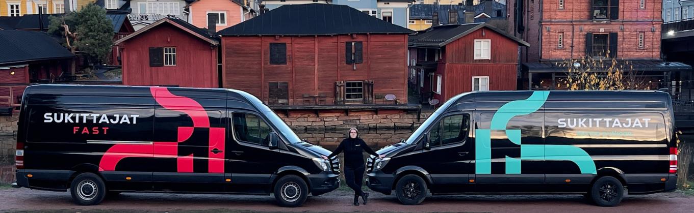 Kaksi mustaa pakettiautoa nokat vastakkain. Autojen välissä seisoo Mervi Svensk. Autoissa lukee Sukittajat