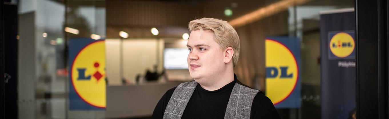 Samu Härkönen seisoo Lidlin pääkonttorin aulassa. Samu katsoo vasemmalle ja hänen taustallaan näkyvät Lildl-kyltit.