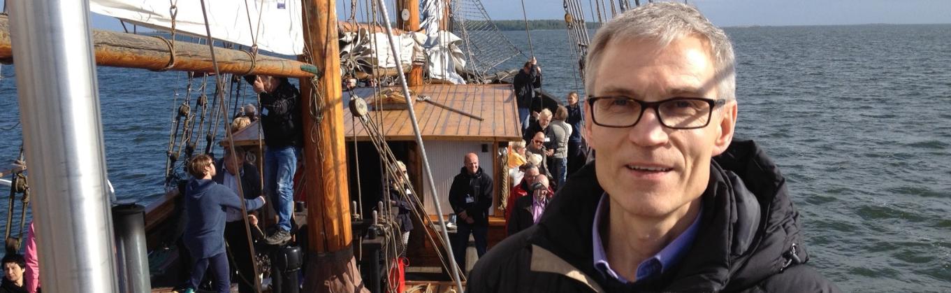 Matti Vesalainen puupurjeveneen kannella. Matilla on harmaat hiukset ja ruskea kevyttoppatakki. Matti hymyilee