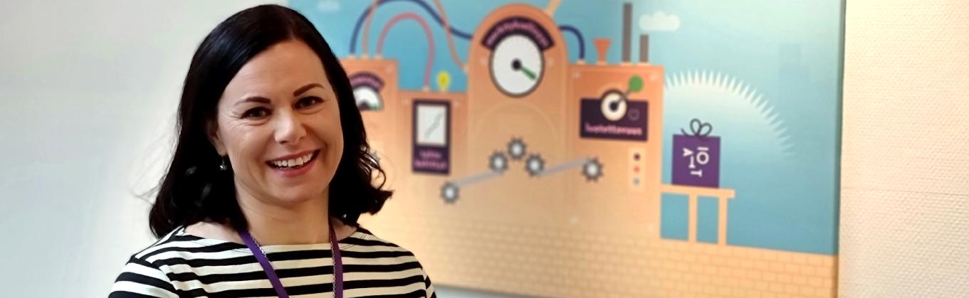 Minna Pohjola hymyilee Taitotalon taulun edessä. Taulu on osa Taitotalon strategiakuvitusta, ja siitä ilmenevät Taitotalon arvot, jotka ovat yhteistyö, tahto kehittyä, luotettavuus ja merkityksellisyys