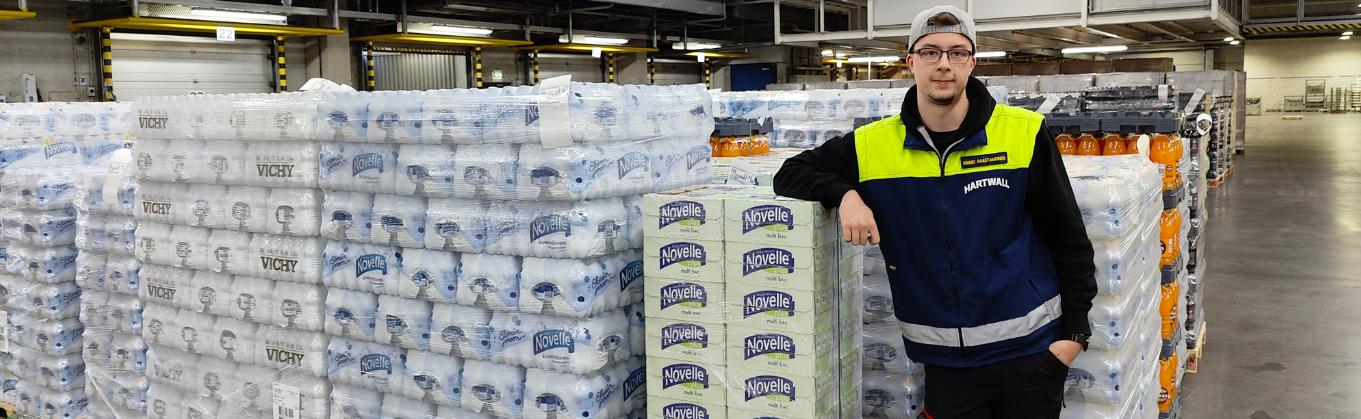 Kimmo Saastamoinen seisoo työpaikallaan Hartwallin varastolla juomalavojen edessä. Lavoilla on erilaisia virvoitusjuomia. Kimmolla on logistiikkatyöntekijän työvaatteet ja hän nojaa oikealla kädellä takana oleviin lavoihin.