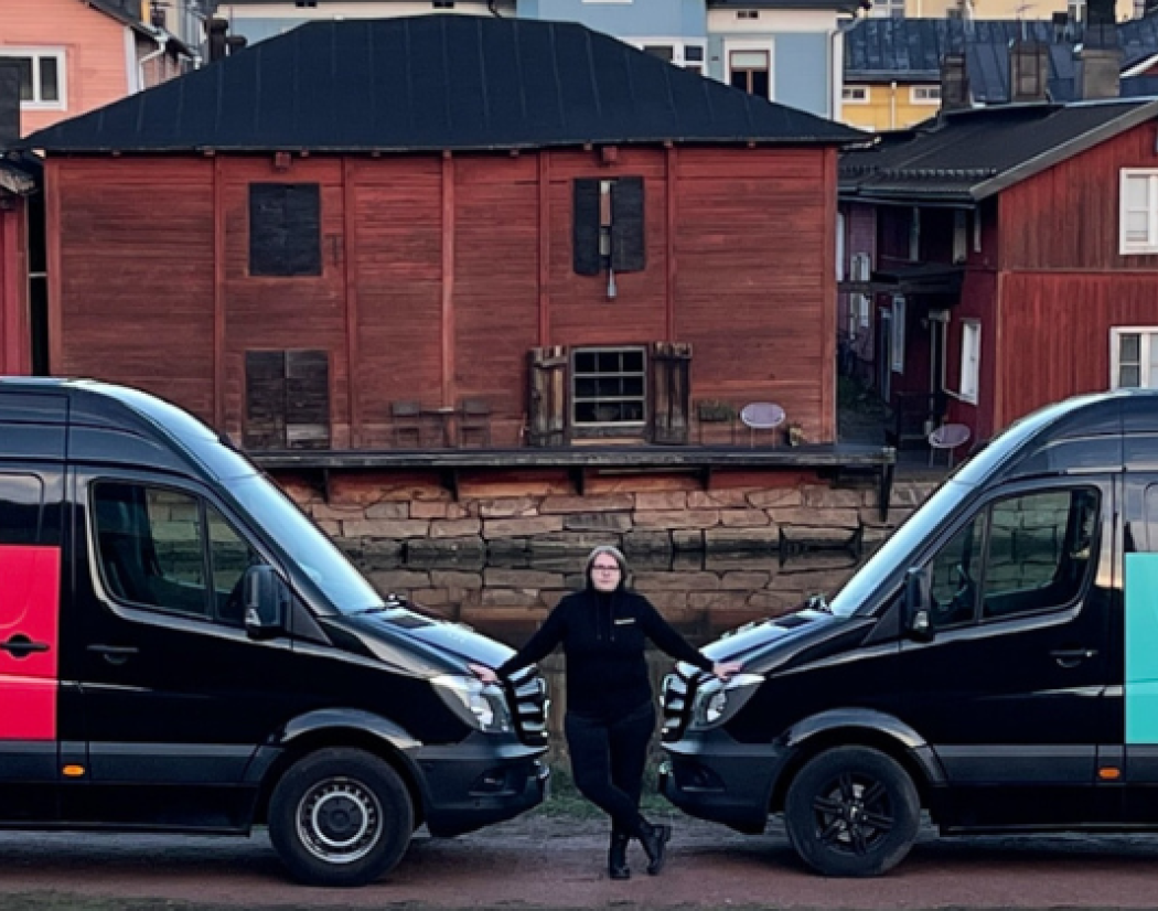 Kaksi mustaa pakettiautoa nokat vastakkain. Autojen välissä seisoo Mervi Svensk. Autoissa lukee Sukittajat