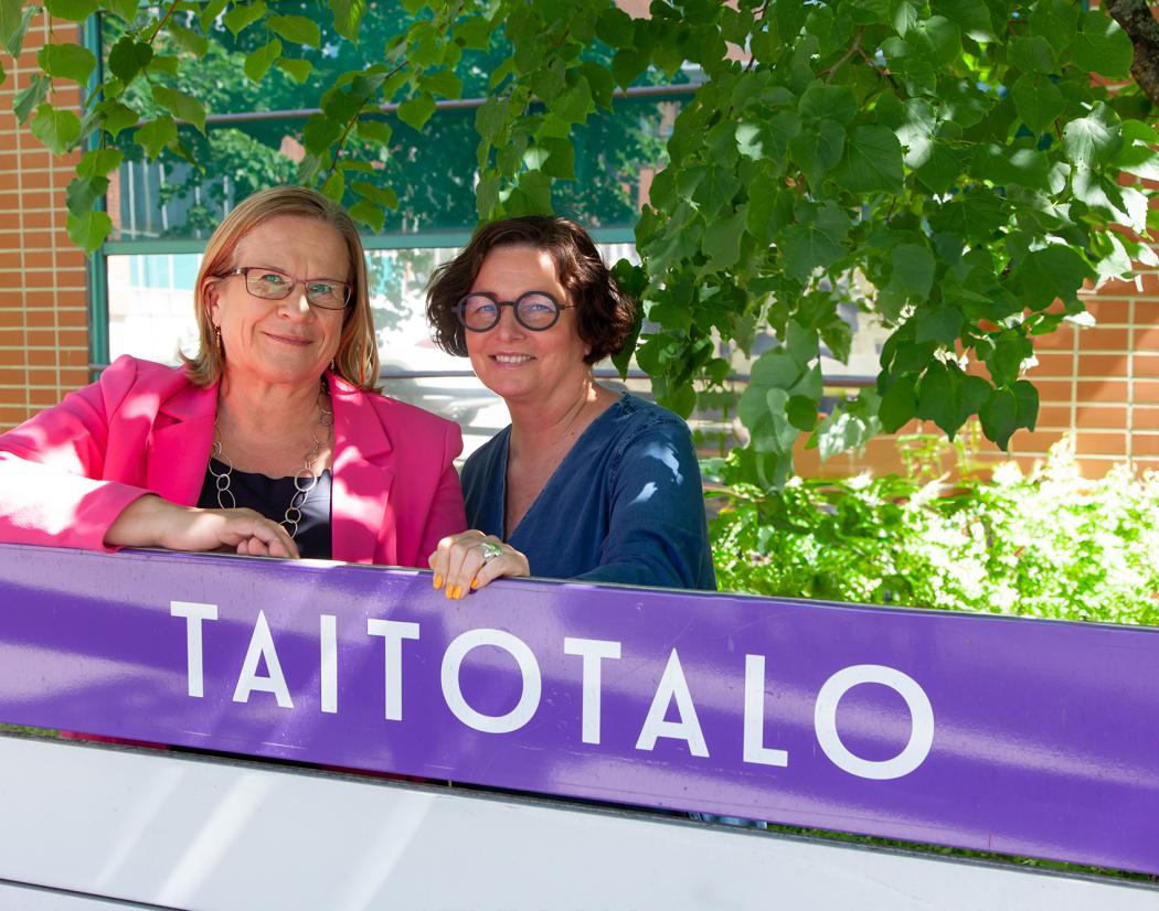 Teija Karjalainen ja Miisa Helenius seisovat kesäistä maisemaa vasten. Heidän edessään on violetti Taitotalo kyltti.