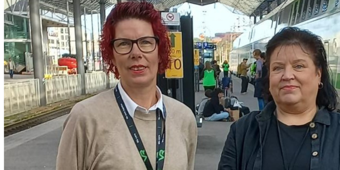 Ann-Mari Ahonen ja Helena Tikka seisovat rautatieasemalla. heidän takanaan on juna ja junaraide.