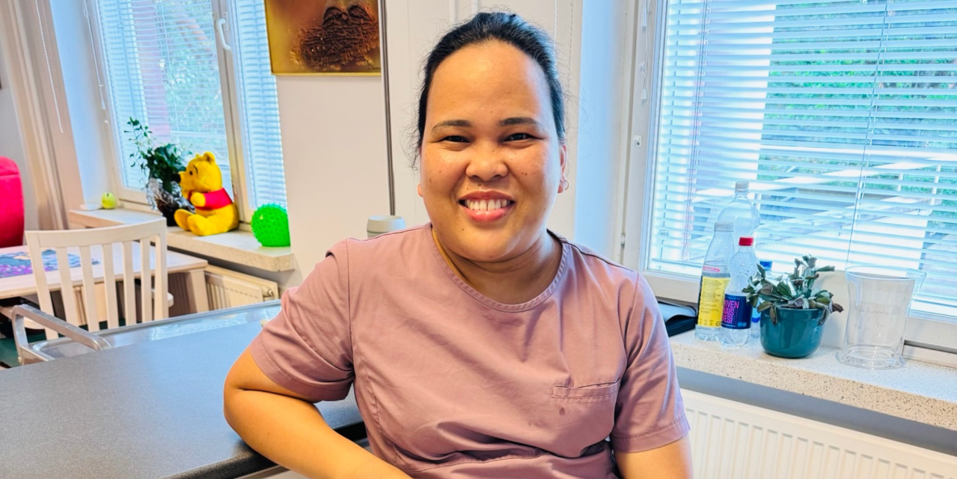Geraldine Arosara työpaikalleen. Geraldine nojaa hoiva-avustajan työasussa pöytään ja katsoo kameraan.