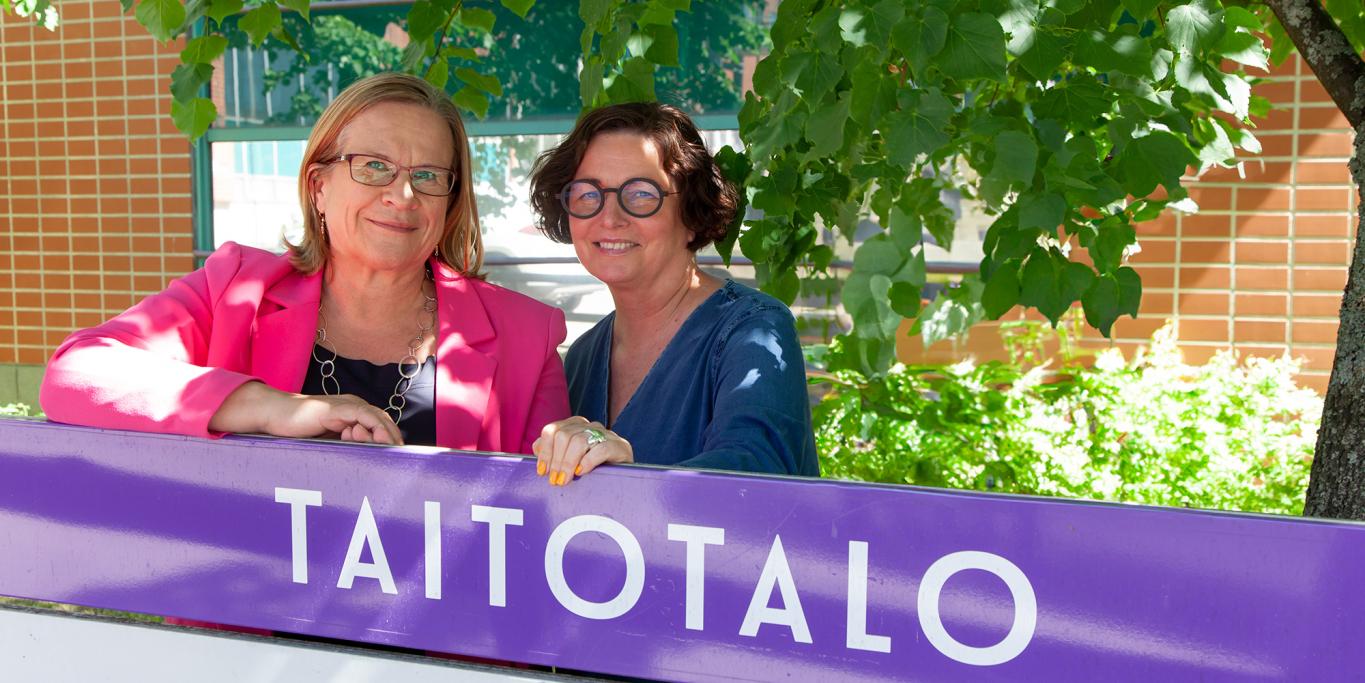 Teija Karjalainen ja Miisa Helenius seisovat kesäistä maisemaa vasten. Heidän edessään on violetti Taitotalo kyltti.