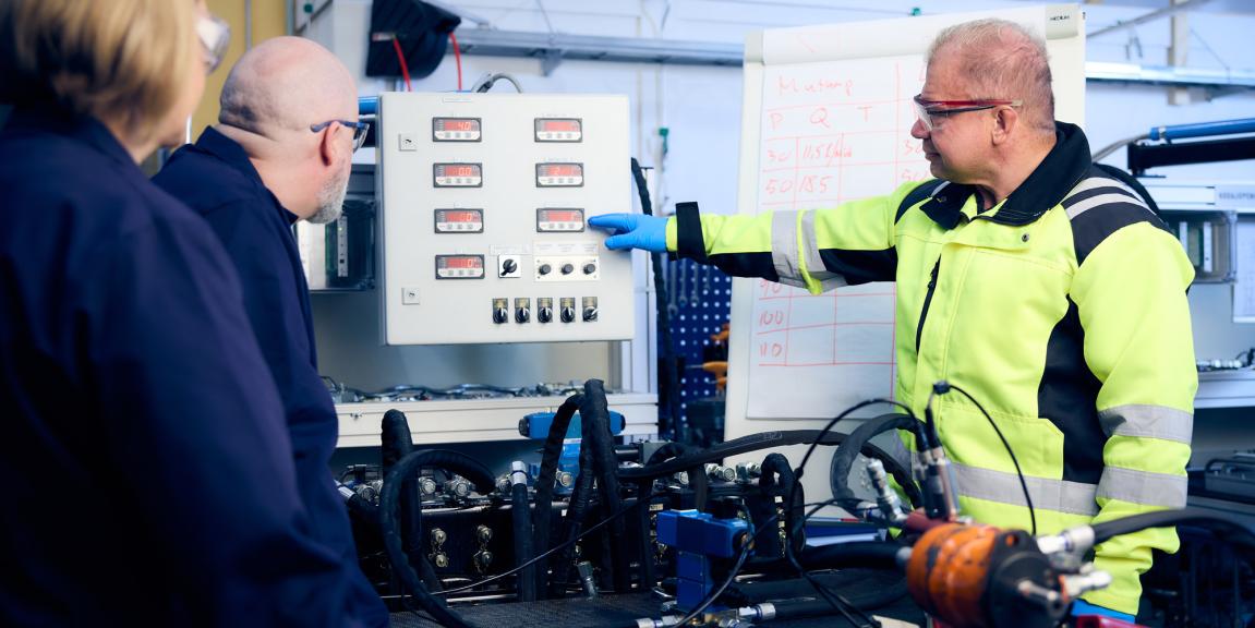 Työtakkinen mies esittää mittaristoa oppilaille hydrauliikkatyötilassa.