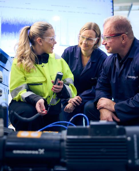 Työtakkinen nainen mittaustilanteessa, kaksi oppilasta katsovat vieressä