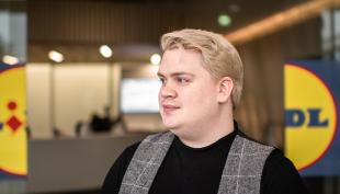 Samu Härkönen seisoo Lidlin pääkonttorin aulassa. Samu katsoo vasemmalle ja hänen taustallaan näkyvät Lildl-kyltit.