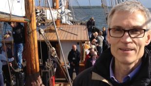 Matti Vesalainen puupurjeveneen kannella. Matilla on harmaat hiukset ja ruskea kevyttoppatakki. Matti hymyilee
