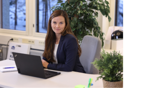 Anne-Mari Mäkinen istuu työpöytänsä äärellä. Hänellä on tietokone edessään, tummat hiukset ja tummansininen jakku. Kuvan toisella puolella on TA-Yhtiöiden logo, joka on sininen teksti TA.FI