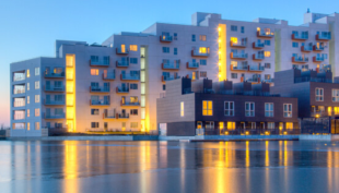 Bravida kuvituskuva, kuvassa kerrostaloja järven rannalla iltahämärässä, auringonvalo kajastaa keltaisena taloja kohti