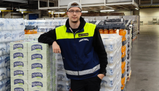 Kimmo Saastamoinen seisoo työpaikallaan Hartwallin varastolla juomalavojen edessä. Lavoilla on erilaisia virvoitusjuomia. Kimmolla on logistiikkatyöntekijän työvaatteet ja hän nojaa oikealla kädellä takana oleviin lavoihin.