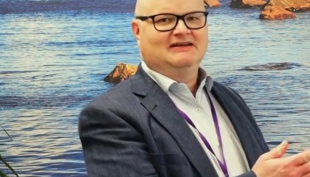 Paso Mustonen on merimaisematapetin edessä. Hänellä on päällään puku ja käsissään tietokone. Kuva rajautuu siten, että kuvassa näkyy vain Pasin yläruumis.a